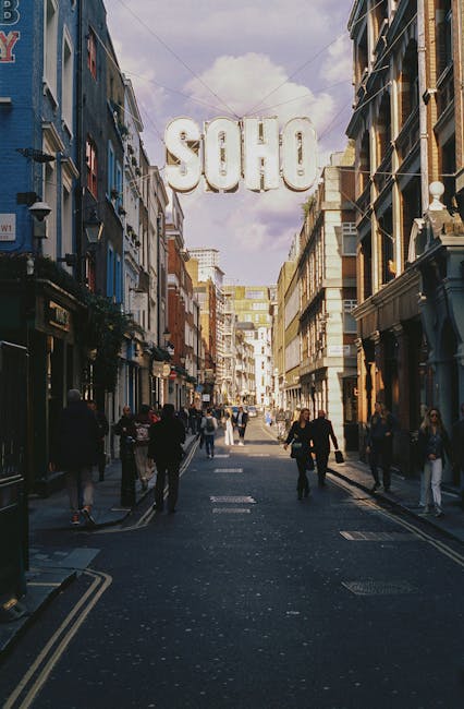 A busy street scene in Soho with pedestrians walking along a wide, paved walkway flanked by multi-storey buildings featuring traditional shop fronts and architectural details. Overhead, a large illuminated sign spells out 'SOHO' in bold, white letters suspended between the buildings, visible against a partly cloudy sky. The street is lined with various retail and hospitality establishments, with some storefronts displaying signage and window displays. The environment suggests an urban area with active foot traffic frequently seen in central London entertainment districts. The scene captures the vibrant atmosphere characteristic of Soho, with the signage indicating the location. The image emphasizes the urban setting and the architecture typical of the area, linking to the context of private waste handling and rubbish removal services suited for bustling commercial districts like Soho, managed by local waste collection specialists such as Rubbish Collection Soho, who handle waste disposal professionally and efficiently.