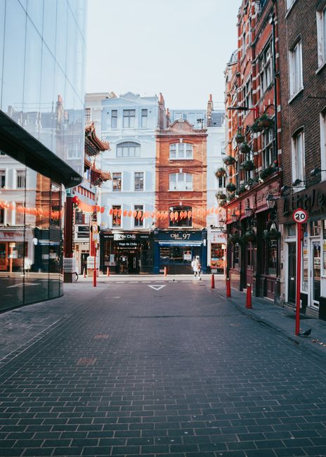 A quiet urban street scene in Soho with a paved pedestrian area flanked by multi-story buildings on both sides. The buildings have a mix of brick and glass facades, with some windows adorned by flower boxes and small decorative elements. In the background, there is a row of older, ornate buildings with varied architectural details. Overhead, red lantern-style string lights are suspended between the buildings, creating a festive atmosphere. The street is lined with short red safety bollards, and a few pedestrians are visible walking nearby. On the right, a shop frontage featuring signage and windows can be seen, while on the left, a modern glass building reflects the surrounding structures. The scene is captured during daylight hours with soft, natural lighting. The image, sourced from Rubbish Collection Soho, suggests an urban environment where independent waste collection or on-site clearance might be necessary to maintain cleanliness in this busy area, especially for local businesses including restaurants and bars.