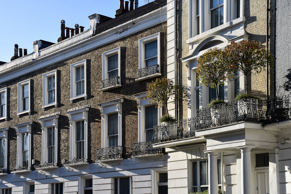 The image depicts a row of historic terraced townhouses with brick facades, typical of urban residential architecture. The building in focus features a light-colored exterior with decorative white mouldings framing the windows and distinguishing the entrance. Each building has multiple sash windows with white frames; some windows are open, revealing interior curtains, and a few have small, ornate iron balcony railings attached beneath them. The balconies are adorned with potted plants and small trees, adding greenery against the brickwork. The street-facing façade is illuminated by natural daylight, casting soft shadows that highlight the architectural details. The rooftops are lined with traditional chimney stacks, some with clay pots. The scene’s overall atmosphere is calm and orderly, embodying a typical cityscape with well-maintained properties. This residential setting could relate to property management or private waste disposal services, where such buildings may require independent rubbish collection. The focus remains on the architectural features, providing a clear context for waste removal in an urban environment, consistent with the service offerings of Rubbish Collection Soho for tailored rubbish removal needs.